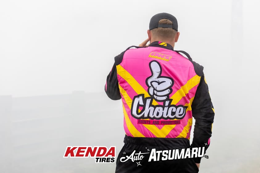 A man viewed from behind wearing a bright pink and yellow racing jacket with a large 'Choice' logo on the back, standing in a foggy outdoor setting. Visible text logos at the bottom include 'Kenda Tires' and 'Atsumaru'.