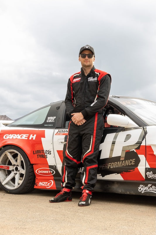 A man in a black, red, and white racing suit stands next to a red, white, and black race car under an overcast sky. The suit has 'Garage H' and 'Drift Co.